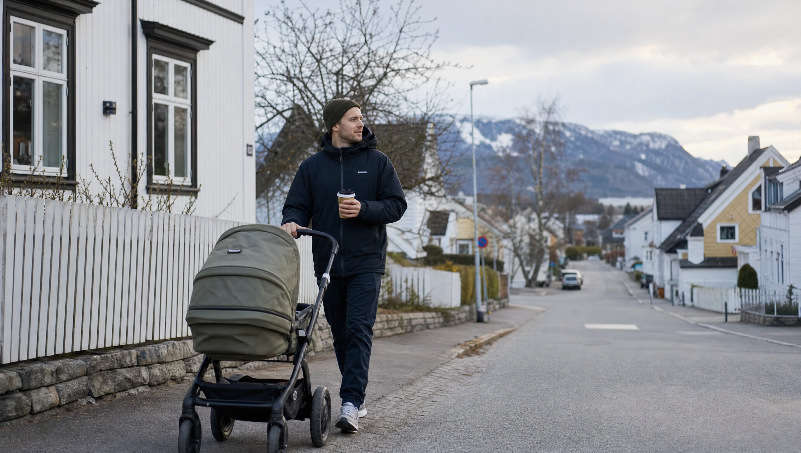 Far går tur med barnevogn på en norsk gate med kaffe i hånden