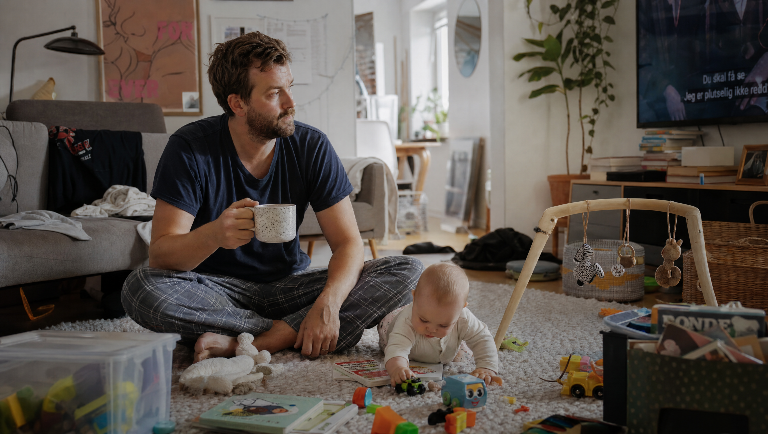 Far hjemme i stua med baby blant leker og en kaffekopp
